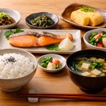 Japanese meal set: grilled salmon on a rectangular plate with lemon, bowls of white rice and miso soup, plus tamagoyaki and assorted vegetables on the side.